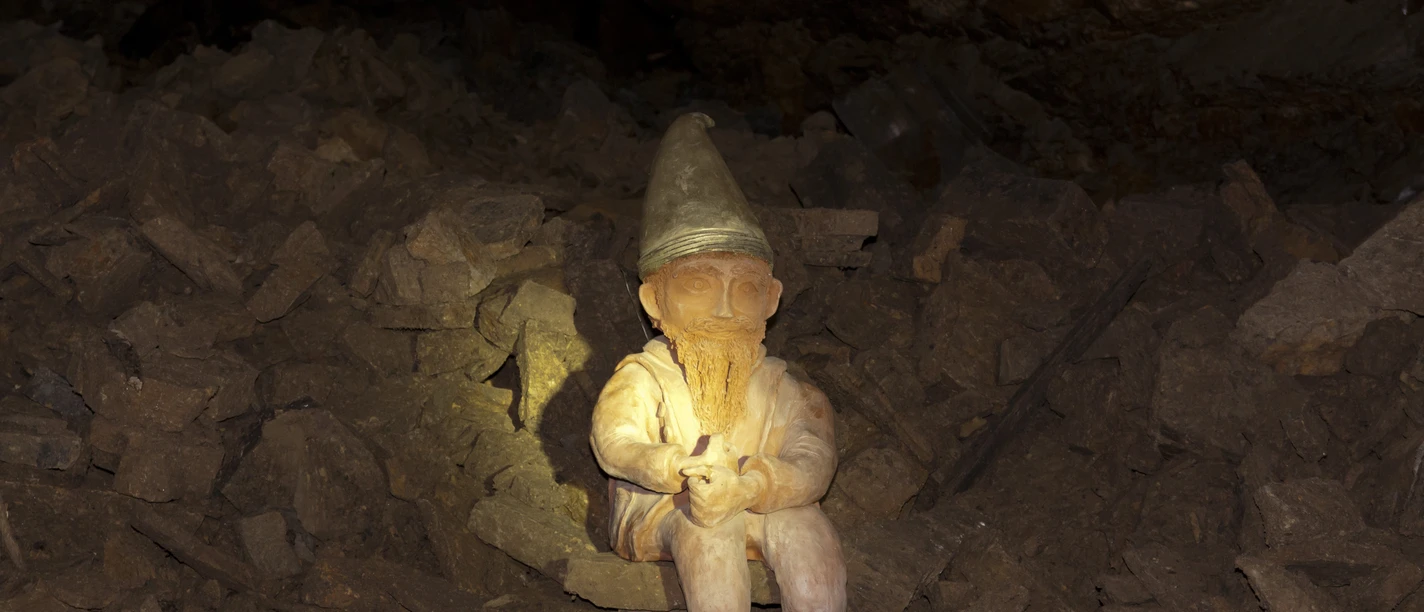 Ein Hüggelzwerg A dwarf with a pointed hat sits on boulders in a dark cave.