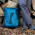 Ein blauer Wanderrucksack und Beine einer Person stehen auf buntem Herbstlaub vor Holzstapeln.