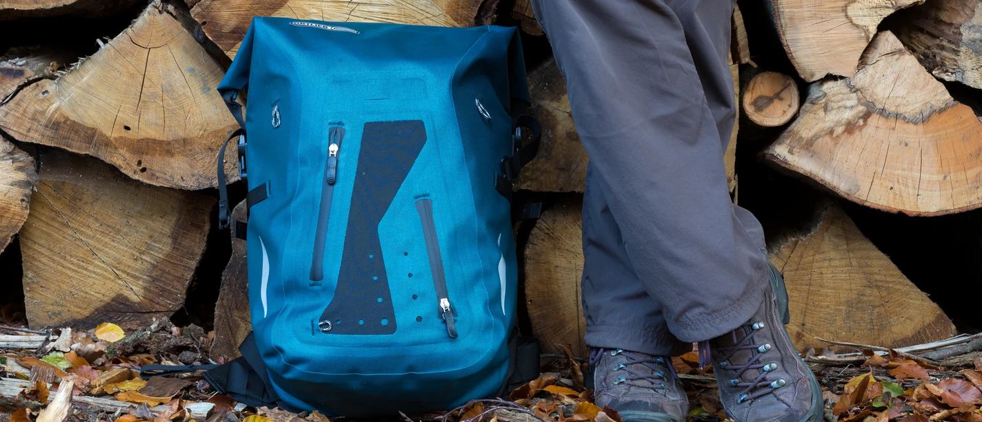 Een blauwe wandelrugzak en de benen van een persoon staan op kleurrijke herfstbladeren voor stapels hout.