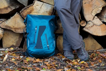 Wanderrast Ein blauer Wanderrucksack und Beine einer Person stehen auf buntem Herbstlaub vor Holzstapeln.