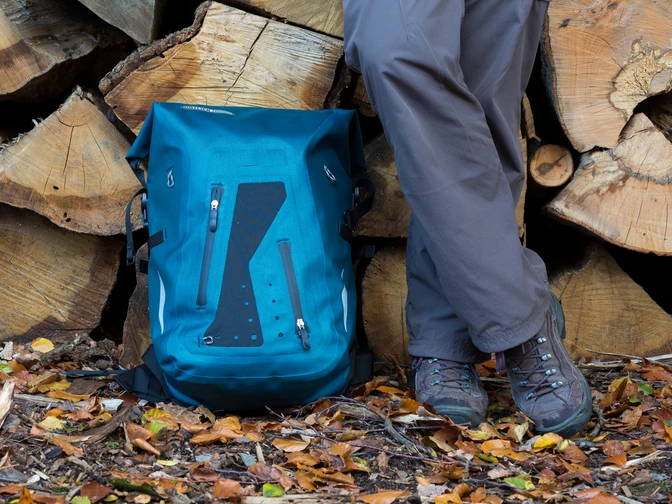 Wanderrast Ein blauer Wanderrucksack und Beine einer Person stehen auf buntem Herbstlaub vor Holzstapeln.A blue hiking rucksack and the legs of a person stand on colorful autumn leaves in front of piles of wood.En blå vandrerygsæk og benene på en person står på farverige efterårsblade foran bunker af træ.Een blauwe wandelrugzak en de benen van een persoon staan op kleurrijke herfstbladeren voor stapels hout.