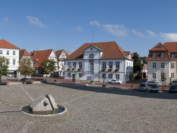 Panorama Marktplatz Quakenbrück.jpg Weitläufiger Platz mit Kopfsteinpflaster, modernen Skulpturen und historischen Gebäuden.