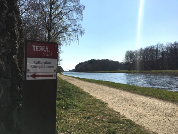 Wandelpad langs het water, omzoomd met bomen in de zon, wegwijzer "Terra.track".