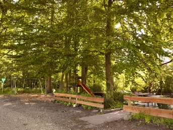 Biergarten Mühlcafé Spielplatz mit Schaukeln und Rutsche unter grünen Laubbäumen, umgeben von Holzzäunen in naturnaher Umgebung.