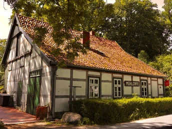 Fachwerkhaus mit rotem Ziegeldach und Holztüren, umgeben von Bäumen, "Mühlcafé" Schild sichtbar.