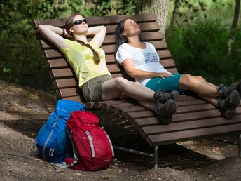 Wellenliege im Nettetal Zwei Wanderinnen entspannen auf einer Holzbank im Schatten eines Waldes mit gepackten Rucksäcken.