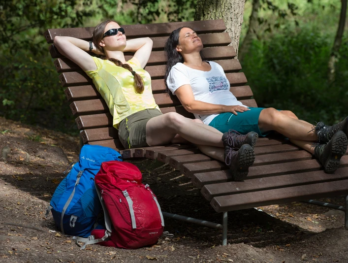 Wellenliege im Nettetal Zwei Wanderinnen entspannen auf einer Holzbank im Schatten eines Waldes mit gepackten Rucksäcken.Two female hikers relax on a wooden bench in the shade of a forest with their rucksacks packed.To kvindelige vandrere slapper af på en træbænk i skyggen af en skov med deres rygsække pakket.Twee vrouwelijke wandelaars relaxen op een houten bankje in de schaduw van een bos met hun rugzakken ingepakt.