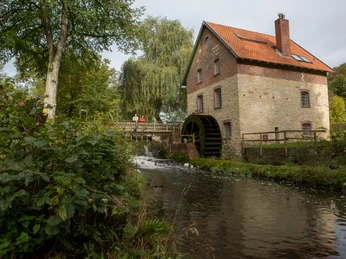 Knollmeyers Mühle Historische Backstein-Wassermühle mit rotem Dach, umgeben von Bäumen und fließendem Bach.Historic brick water mill with a red roof, surrounded by trees and a flowing stream.Historisk vandmølle i mursten med rødt tag, omgivet af træer og et rindende vandløb.Historische bakstenen watermolen met een rood dak, omringd door bomen en een stromend beekje.