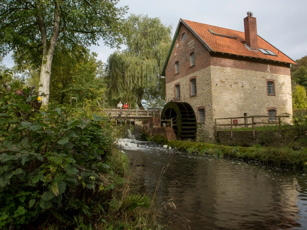 Knollmeyers Mühle Historic brick water mill with a red roof, surrounded by trees and a flowing stream.