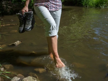 Erfrischung in der Nette Barfuß überquert eine Person einen flachen, klaren Bach mit Wanderschuhen in der Hand.Barefoot, a person crosses a shallow, clear stream with hiking boots in hand.Barfodet krydser en person et lavt, klart vandløb med vandrestøvler i hånden.Op blote voeten steekt iemand een ondiep, helder beekje over met wandelschoenen in de hand.