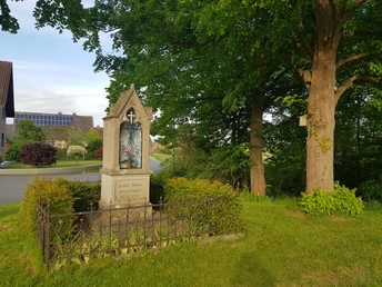Ein historischer Bildstock auf grüner Wiese vor ländlicher Kulisse mit Bäumen und Häusern im Hintergrund.