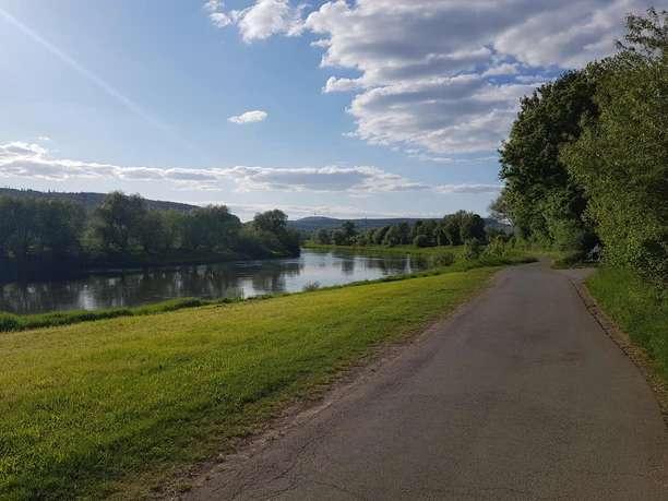 1_weseraue_abzweigholzminden_hx2_stephanberg Ein ruhiger Fluss schlängelt sich durch eine grüne Landschaft, flankiert von Bäumen und einem blauen Himmel.