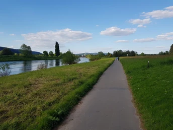 2_radweg_blickaufholzminden_hx2_stephanberg Fahrradweg entlang eines Flusses, umgeben von grüner Landschaft, mit zwei Personen im Hintergrund.