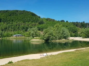 Grüne Hügel und ein klarer See mit Sandstrand, umgeben von Bäumen unter blauem Himmel.