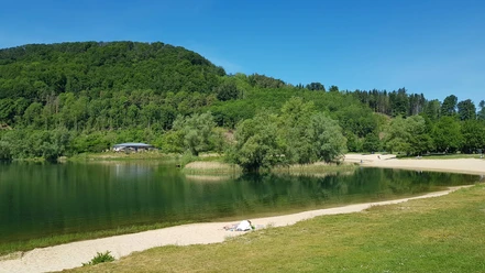 1_freizeitanlage_hx3_stephanberg Grüne Hügel und ein klarer See mit Sandstrand, umgeben von Bäumen unter blauem Himmel.
