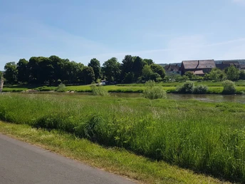2_blickaufboffzen_hx3_stephanberg Der Blick zeigt eine grüne Flusslandschaft mit einem Dorf im Hintergrund unter blauem Himmel.