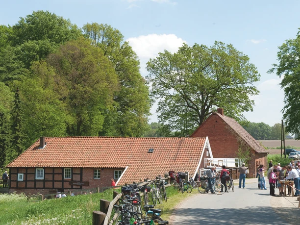 Wöstenesch Wassermühle Historisches Fachwerkhaus mit rotem Ziegeldach, umgeben von Bäumen und Menschen mit Fahrrädern.