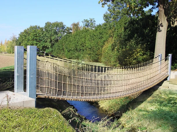 Hängebrücke Rundwanderweg Benkhausen Hängebrücke aus Holz und Seilen über einen kleinen Bach, umgeben von Bäumen und Wiesen.
