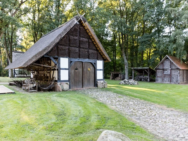 Museumshof Rahden Reetgedecktes Fachwerkhaus mit geschichtsträchtigen landwirtschaftlichen Geräten, umgeben von Bäumen.