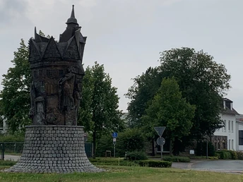 Ein bronzenes Turmdenkmal steht auf einem Kreisverkehr, umgeben von Bäumen und einer Straße.