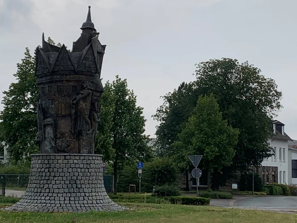 Ein bronzenes Turmdenkmal steht auf einem Kreisverkehr, umgeben von Bäumen und einer Straße.