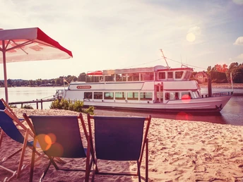 Ein Ausflugsschiff liegt an einem sonnigen Sandstrand, gesäumt von Liegestühlen und einem Sonnenschirm.
