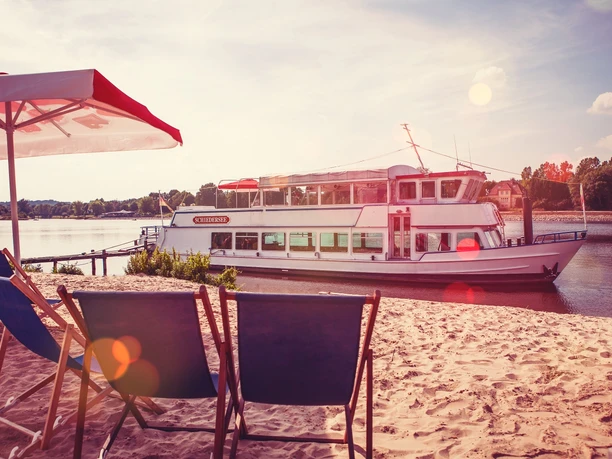 Ein Ausflugsschiff liegt an einem sonnigen Sandstrand, gesäumt von Liegestühlen und einem Sonnenschirm.