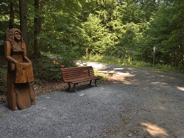 Sterntaler Holzskulptur einer Frau neben einer Parkbank in einem sonnendurchfluteten Waldweg mit Bäumen im Hintergrund.
