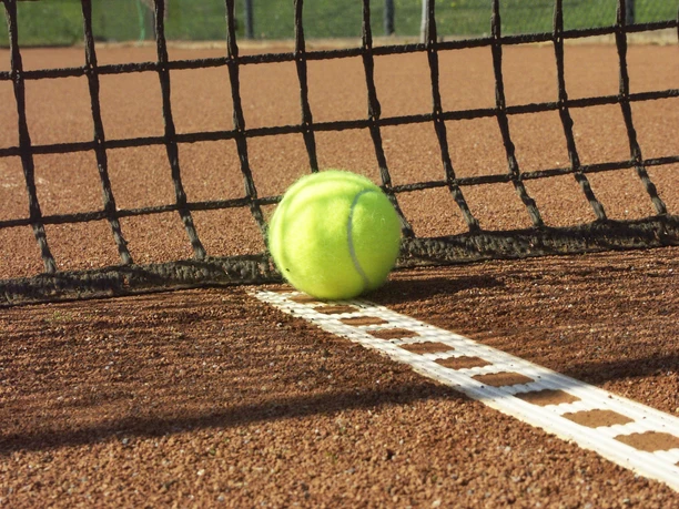 Tennis Ein gelber Tennisball liegt auf der weißen Linie eines roten Sandplatzes direkt am schwarzen Netz.