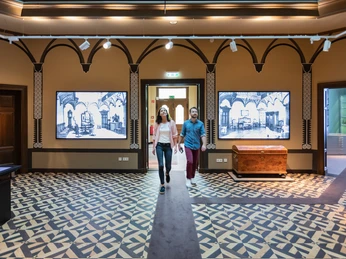 Zwei Personen schlendern durch ein Museum mit historischen Wandtafeln und kunstvollem Bodenmuster.
