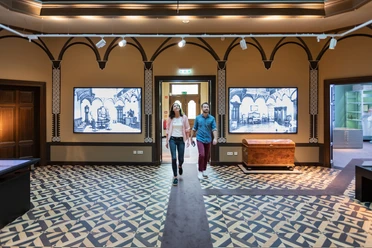 herford-daniel-poeppelmann-haus-teutoburger-wald-tourismus-m-schoberer-2 Zwei Personen schlendern durch ein Museum mit historischen Wandtafeln und kunstvollem Bodenmuster.