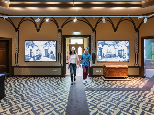herford-daniel-poeppelmann-haus-teutoburger-wald-tourismus-m-schoberer-2 Zwei Personen schlendern durch ein Museum mit historischen Wandtafeln und kunstvollem Bodenmuster.