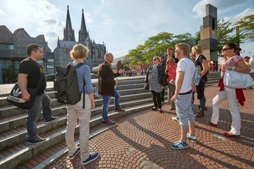A group of people stand on a staircase in Cologne. The striking Cologne Cathedral and modern architecture can be seen in the background. A tour guide gestures during a city tour in the sunshine, surrounded by interested tourists exploring the history and culture of Cologne.