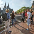 Eine Gruppe von Menschen steht auf einer Treppe in Köln. Im Hintergrund sind der markante Kölner Dom und moderne Architektur zu sehen. Ein Reiseleiter gestikuliert während einer Stadtführung im Sonnenschein, umgeben von interessierten Tourist*innen, die die Geschichte und Kultur Kölns erkunden.A group of people stand on a staircase in Cologne. The striking Cologne Cathedral and modern architecture can be seen in the background. A tour guide gestures during a city tour in the sunshine, surrounded by interested tourists exploring the history and culture of Cologne.