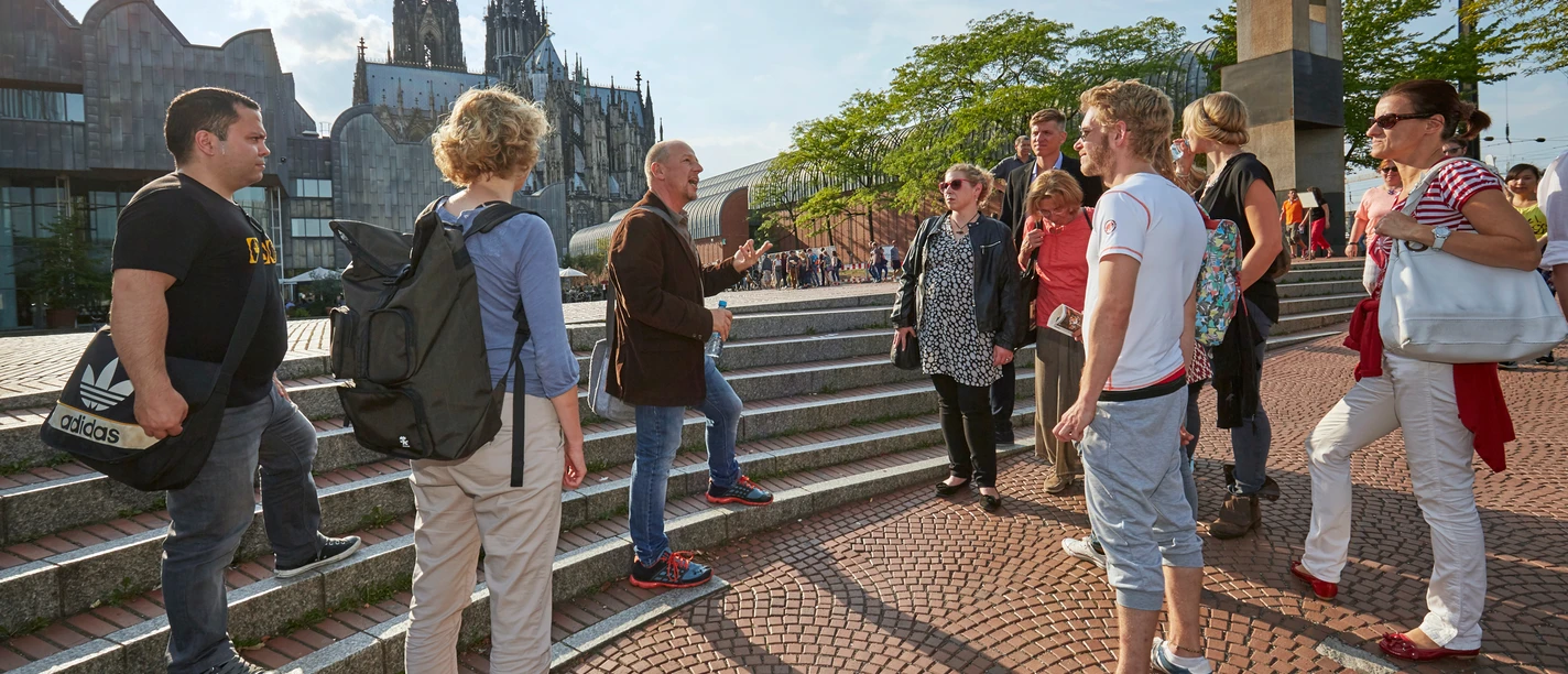 A group of people stand on a staircase in Cologne. The striking Cologne Cathedral and modern architecture can be seen in the background. A tour guide gestures during a city tour in the sunshine, surrounded by interested tourists exploring the history and culture of Cologne.