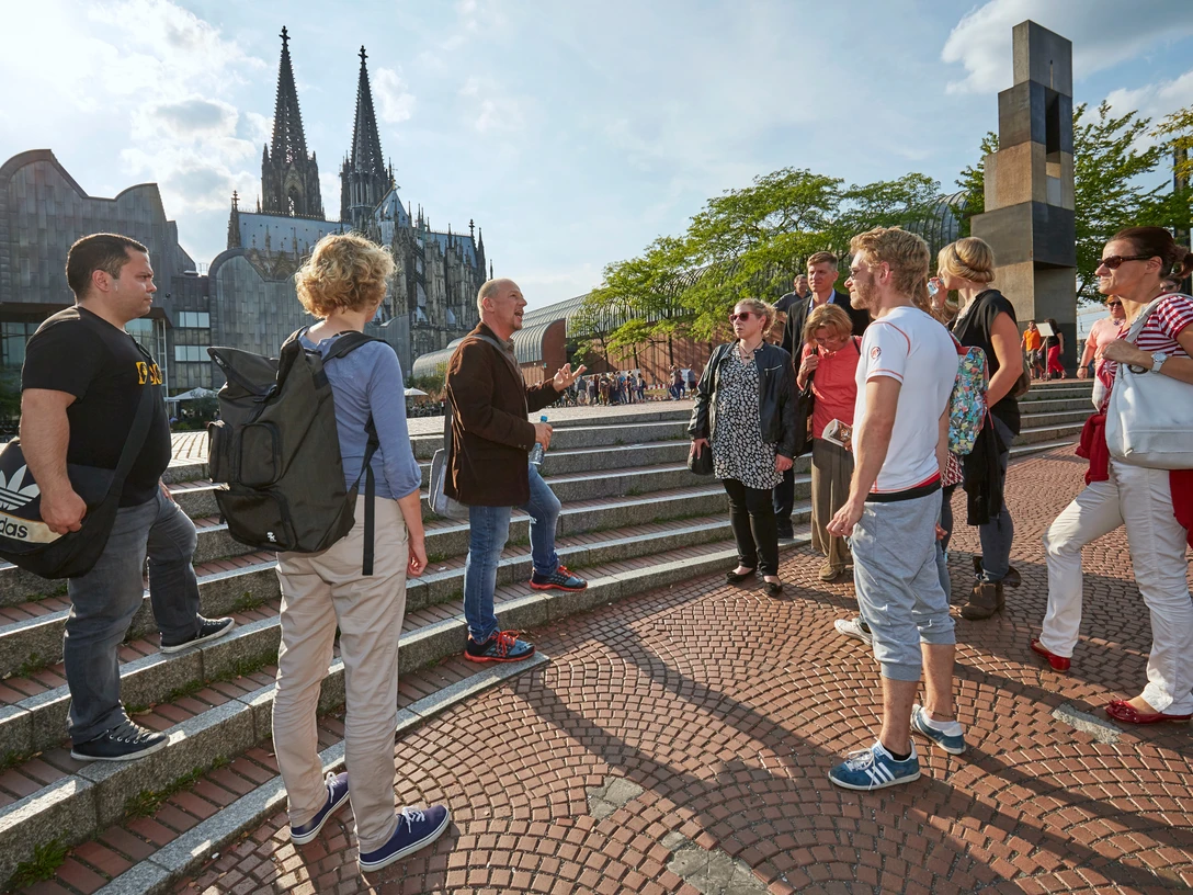 stadtfuehrung-in-koeln-dieter-jacobi-koelntourismus-gmbh Eine Gruppe von Menschen steht auf einer Treppe in Köln. Im Hintergrund sind der markante Kölner Dom und moderne Architektur zu sehen. Ein Reiseleiter gestikuliert während einer Stadtführung im Sonnenschein, umgeben von interessierten Tourist*innen, die die Geschichte und Kultur Kölns erkunden.A group of people stand on a staircase in Cologne. The striking Cologne Cathedral and modern architecture can be seen in the background. A tour guide gestures during a city tour in the sunshine, surrounded by interested tourists exploring the history and culture of Cologne.