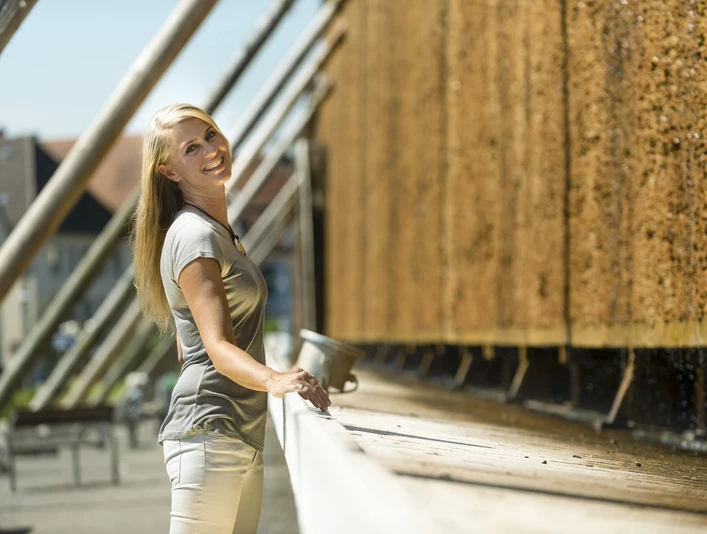 Tief durchatmen am Gradierwerk Eine Frau lehnt lächelnd an einem hölzernen Gradierwerk, das mit herabfließender Sole benetzt ist.