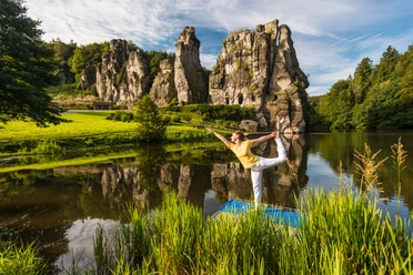 Yoga vor den Externsteinen Eine Person praktiziert Yoga auf einer Plattform vor den majestätischen Externsteinen in einer grünen Umgebung.