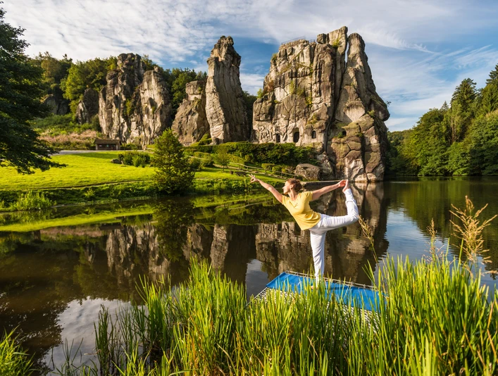 Yoga vor den Externsteinen Eine Person praktiziert Yoga auf einer Plattform vor den majestätischen Externsteinen in einer grünen Umgebung.