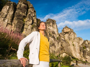 Entspannung in therapeutischer Landschaft Frau lehnt sich entspannt an ein Holzgeländer vor monumentalen Felsformationen unter blauem Himmel.