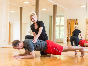 bad-driburg-rueckenuebung-ketz Ein Mann in roter Sporthose macht eine Planke, während eine Trainerin ihn in einem hellen Raum anleitet.