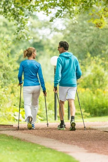 Zwei Personen in sportlicher Kleidung walken mit Stöcken auf einem Parkweg, umgeben von Bäumen.