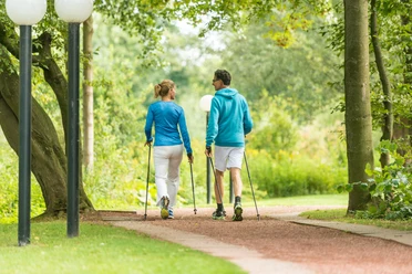 In Bewegung bleiben mit Nordic Walking Zwei Personen in sportlicher Kleidung walken mit Stöcken auf einem Parkweg, umgeben von Bäumen.