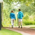 In Bewegung bleiben mit Nordic Walking Zwei Personen in sportlicher Kleidung walken mit Stöcken auf einem Parkweg, umgeben von Bäumen.