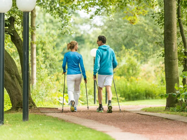 In Bewegung bleiben mit Nordic Walking Zwei Personen in sportlicher Kleidung walken mit Stöcken auf einem Parkweg, umgeben von Bäumen.
