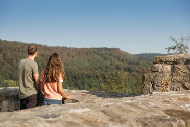 detmold-ruine-falkenburg-teutoburger-wald-tourismus-m-schoberer-4 Zwei Menschen genießen von einer Mauer am Berg aus den Blick auf die bewaldete Landschaft.