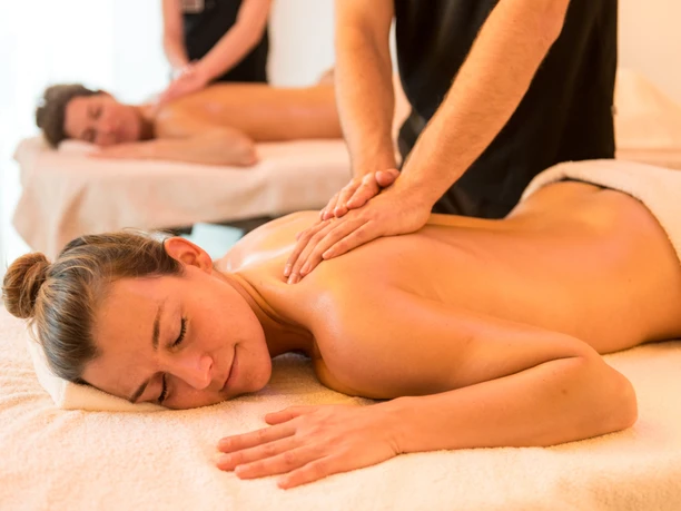 Zwei Personen genießen entspannende Rückenmassagen auf Massageliegen in einem Wellnessbereich.