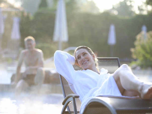 Frau in weißem Bademantel entspannt auf einer Liege am Pool, während ein Mann im Wasser ist.