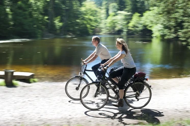 Radfahrer Zwei Menschen radeln entlang eines Flusses inmitten eines bewaldeten Gebiets bei sonnigem Wetter.