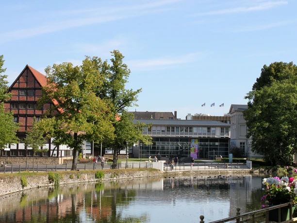 Historisches Fachwerkgebäude neben modernem Museumsbau am Wasser, umgeben von grünen Bäumen.